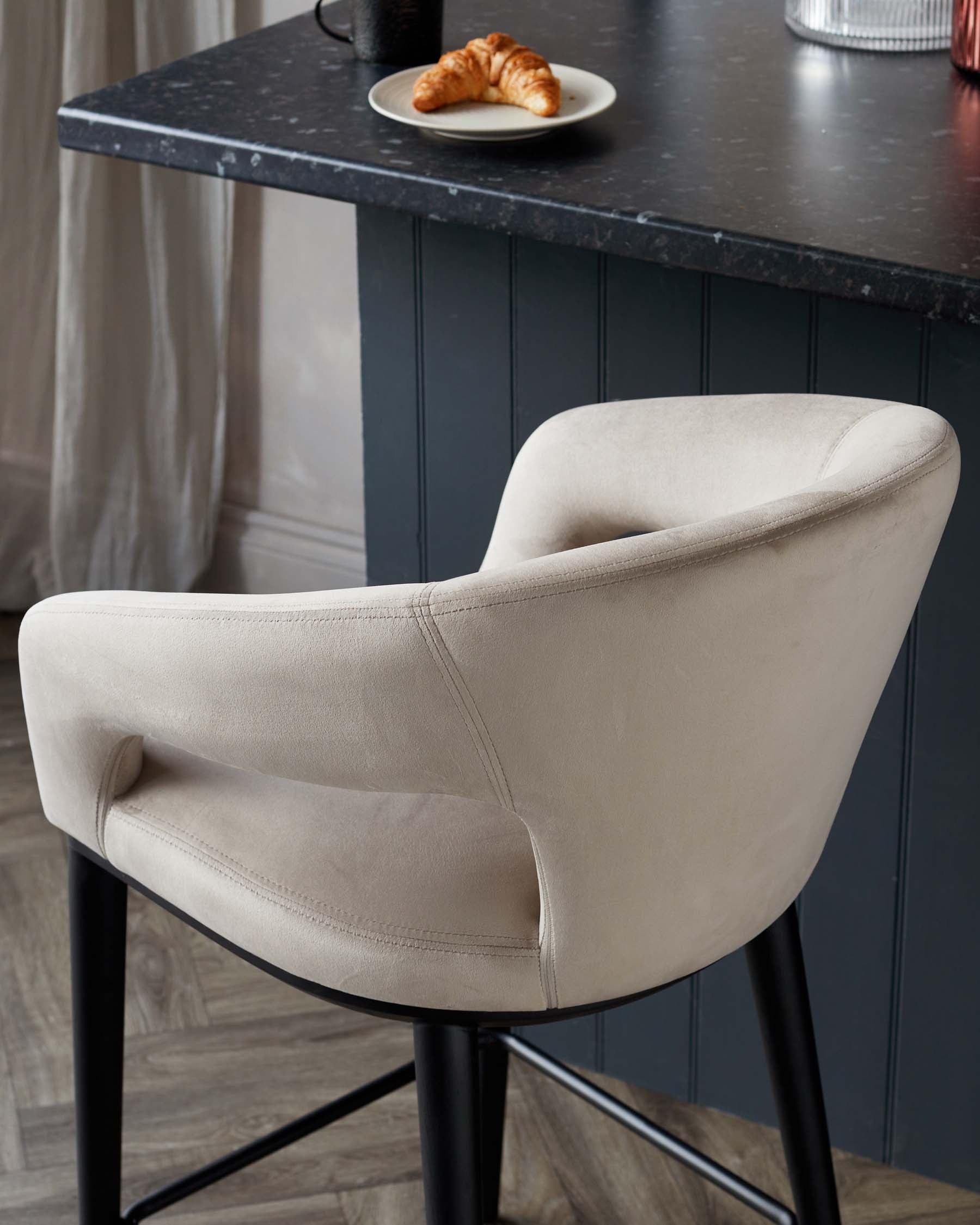 Modern light beige upholstered dining chair with a curved backrest and armrests, featuring an elegant black metal base. Positioned next to a dark stone table with a plate of croissants.