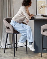 Elegant modern bar stool with a smooth taupe upholstery, featuring a high backrest and armrests. The stool has slender black metal legs with a geometrical footrest design, providing a stylish and comfortable seating option for contemporary high-table settings.