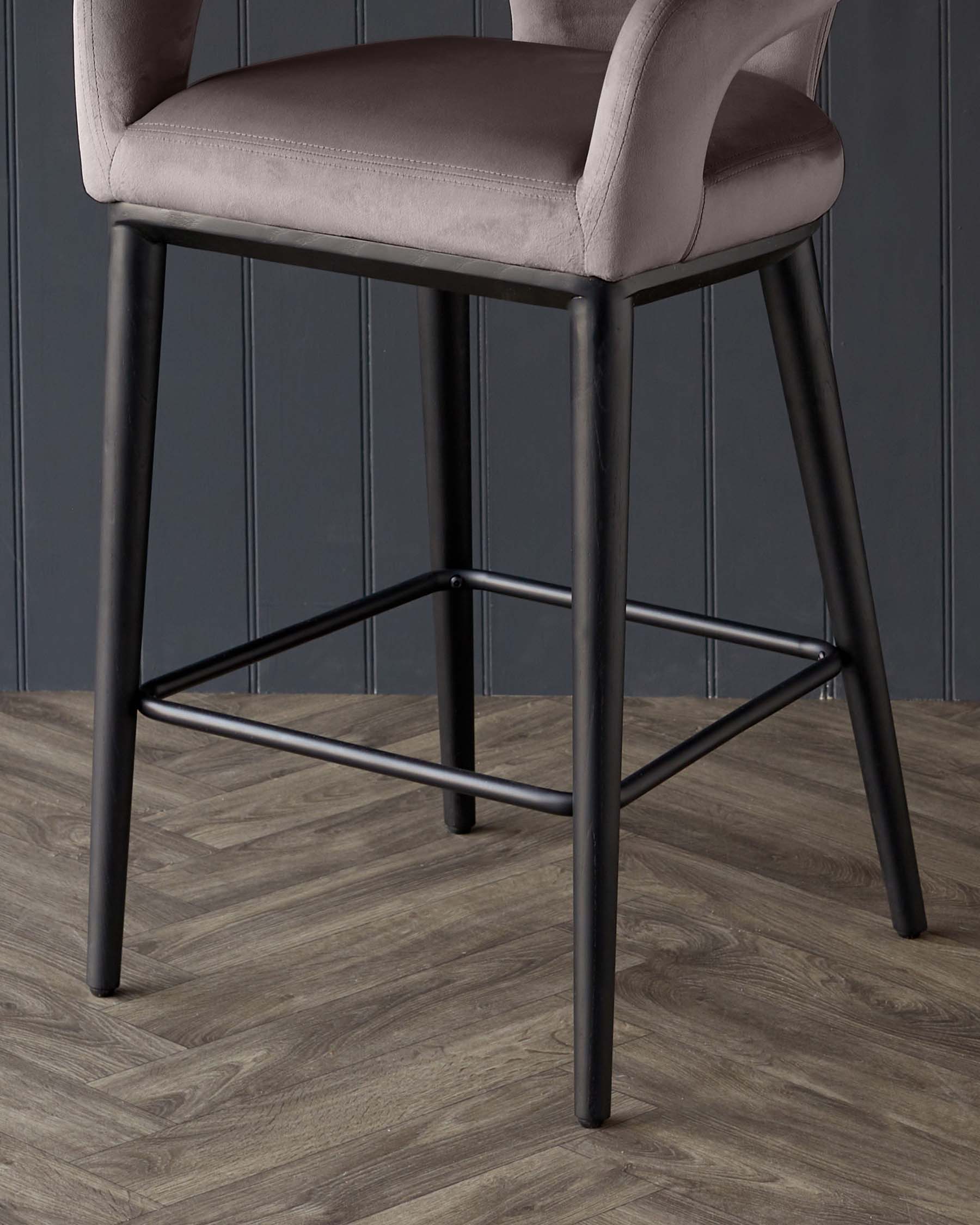 Modern bar stool with durable black metal legs and a built-in footrest, featuring a plush seat and backrest upholstered in soft mauve fabric, against a dark panelled wall and wooden floor.