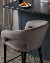 Elegant modern bar chair with a curved backrest and armrests, upholstered in taupe fabric, paired with slim black metal legs, positioned by a dark counter with a plate of croissants.