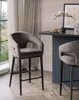 Modern upholstered bar stools with curved backrests and slim, black metal legs, displayed alongside a dark kitchen counter.