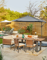 Outdoor dining set featuring a rectangular wooden table, cushioned corner sofa, and rattan-back chairs under a grey umbrella.