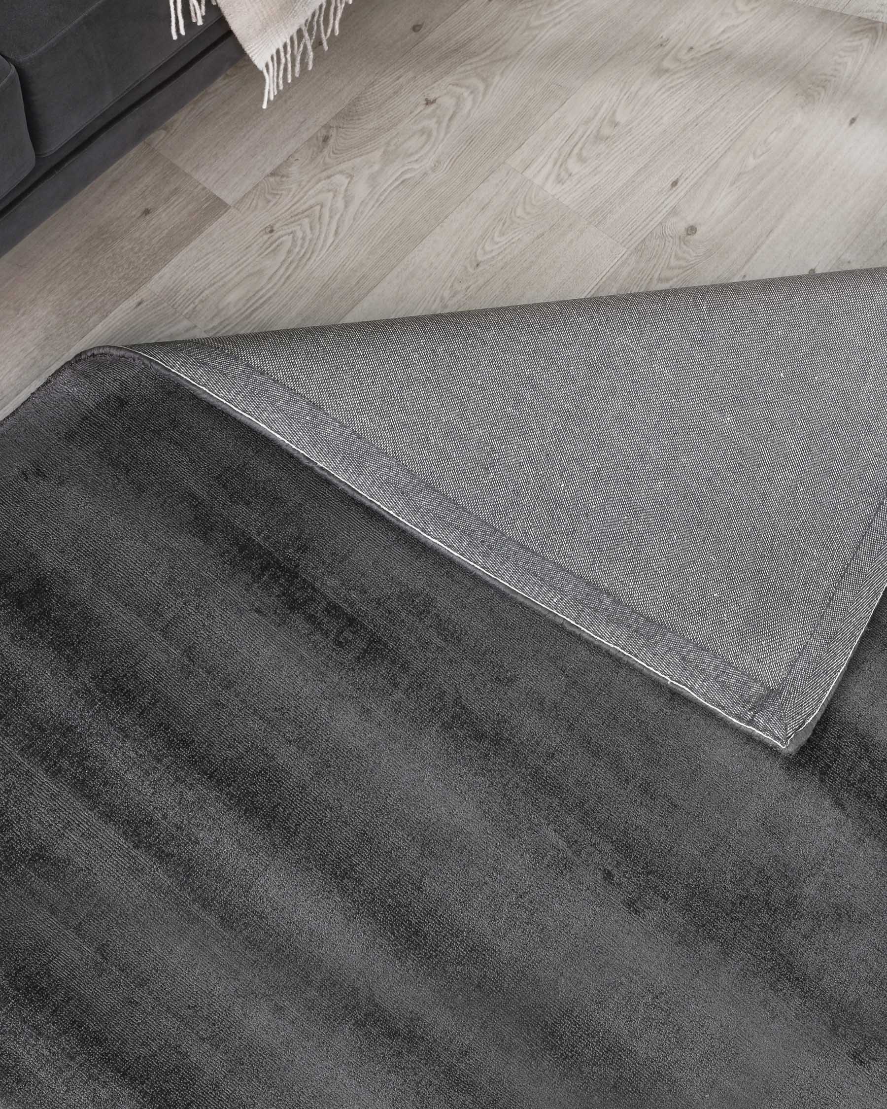 Elegant textured area rug in shades of dark grey with a lighter grey border, partially visible on a light wooden floor beside a black or dark-toned couch with a fringed throw blanket casually draped over it.