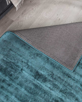 Plush teal area rug with a textured pattern and neatly bordered edges overlaid by a corner of a light grey woven throw blanket on a wooden floor.