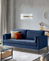 Elegant navy blue three-seater sofa with a plush velvet finish, featuring clean lines and minimalist metal legs. Sofa is accessorized with two beige and one orange decorative pillows, paired with a contemporary rectangular glass-top coffee table with a black metal frame and wooden shelf.