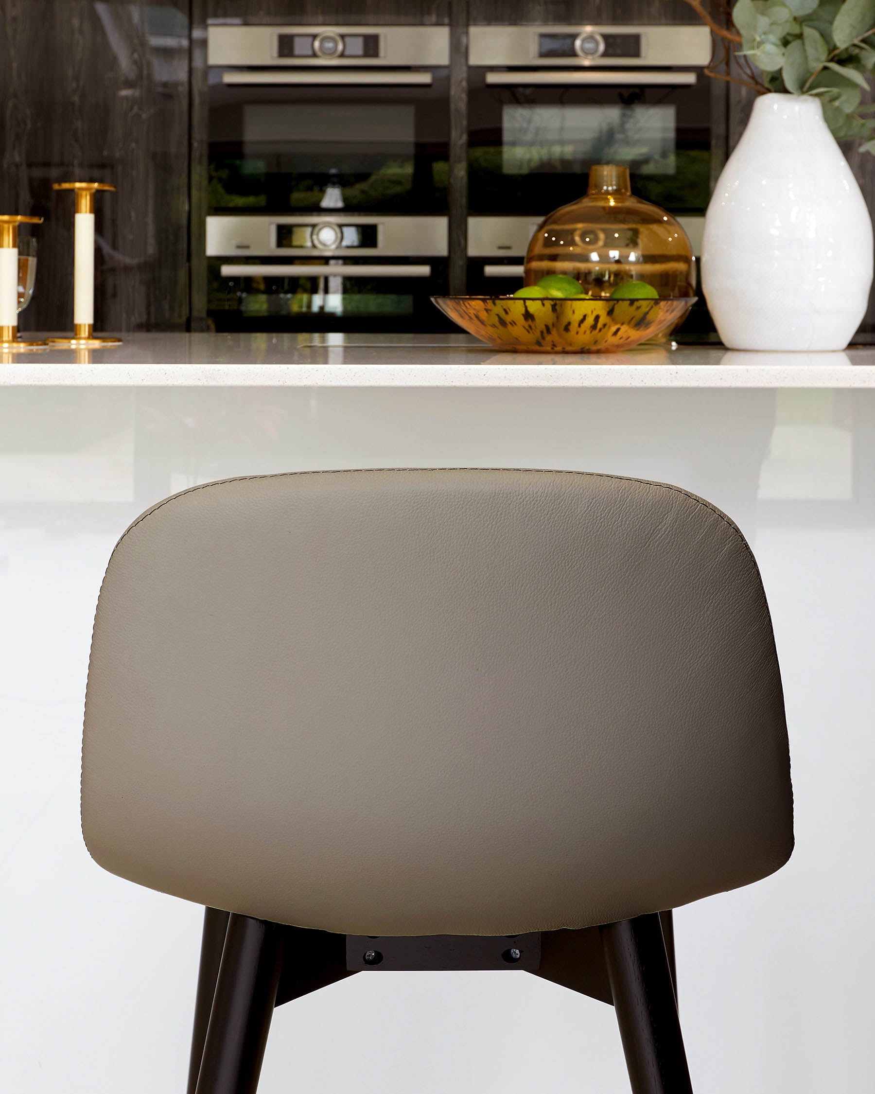 Modern taupe counter stool with a curved backrest and angular black legs, set against a kitchen island with a white countertop and sophisticated cabinetry. Decorative elements include a brown glass vase and a white ceramic vase on the countertop.