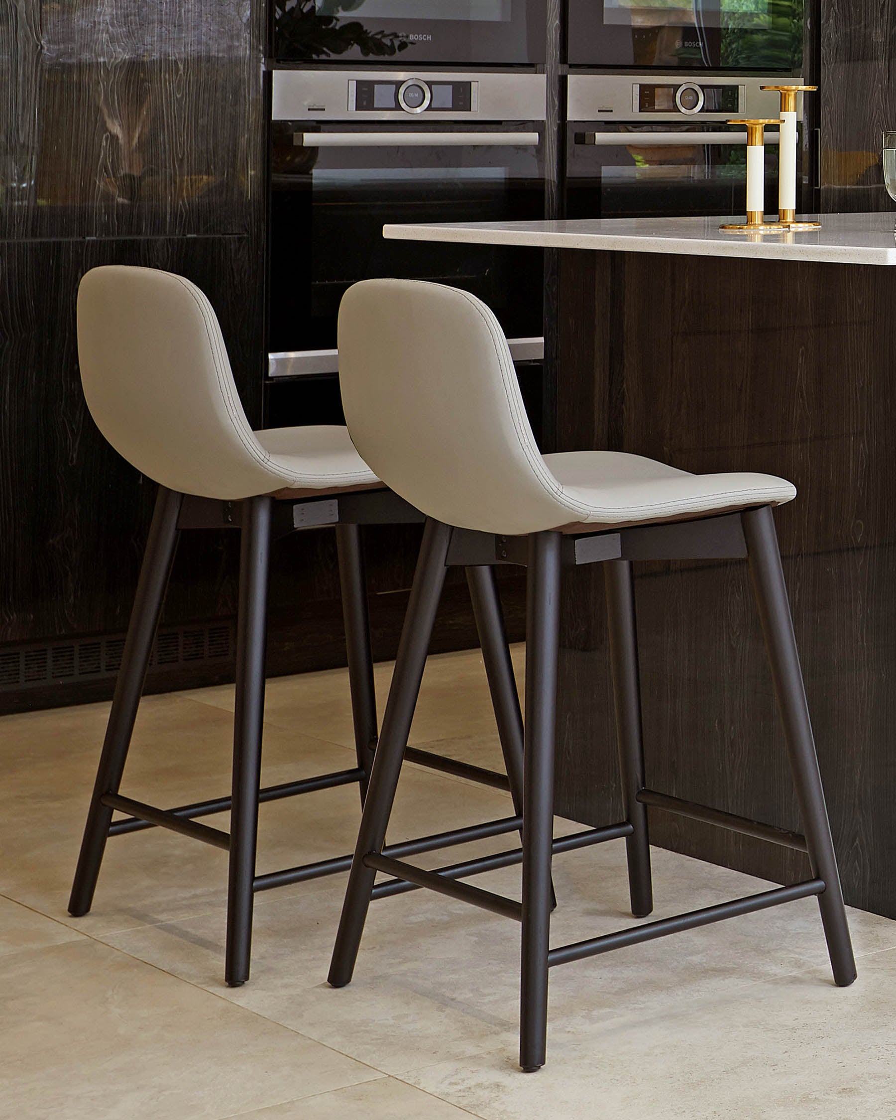 Modern-style kitchen bar stools with sleek taupe upholstery on the seat and back, and a dark brown, almost black, metal frame with four slender legs and footrests.