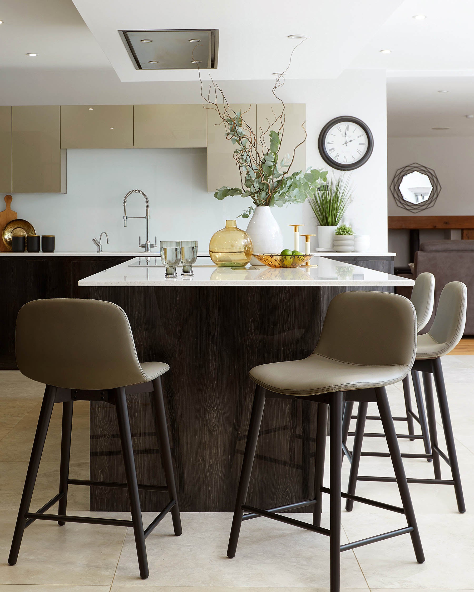 Three modern bar stools with dark wooden legs and a taupe, leather-upholstered seat and backrest, positioned around a kitchen island with a dark wood finish and a white countertop.