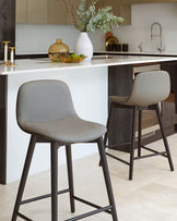 Two modern grey bar stools with sleek, ergonomic designs, featuring curved backrests and angled metal legs, positioned at a kitchen island with a white countertop.