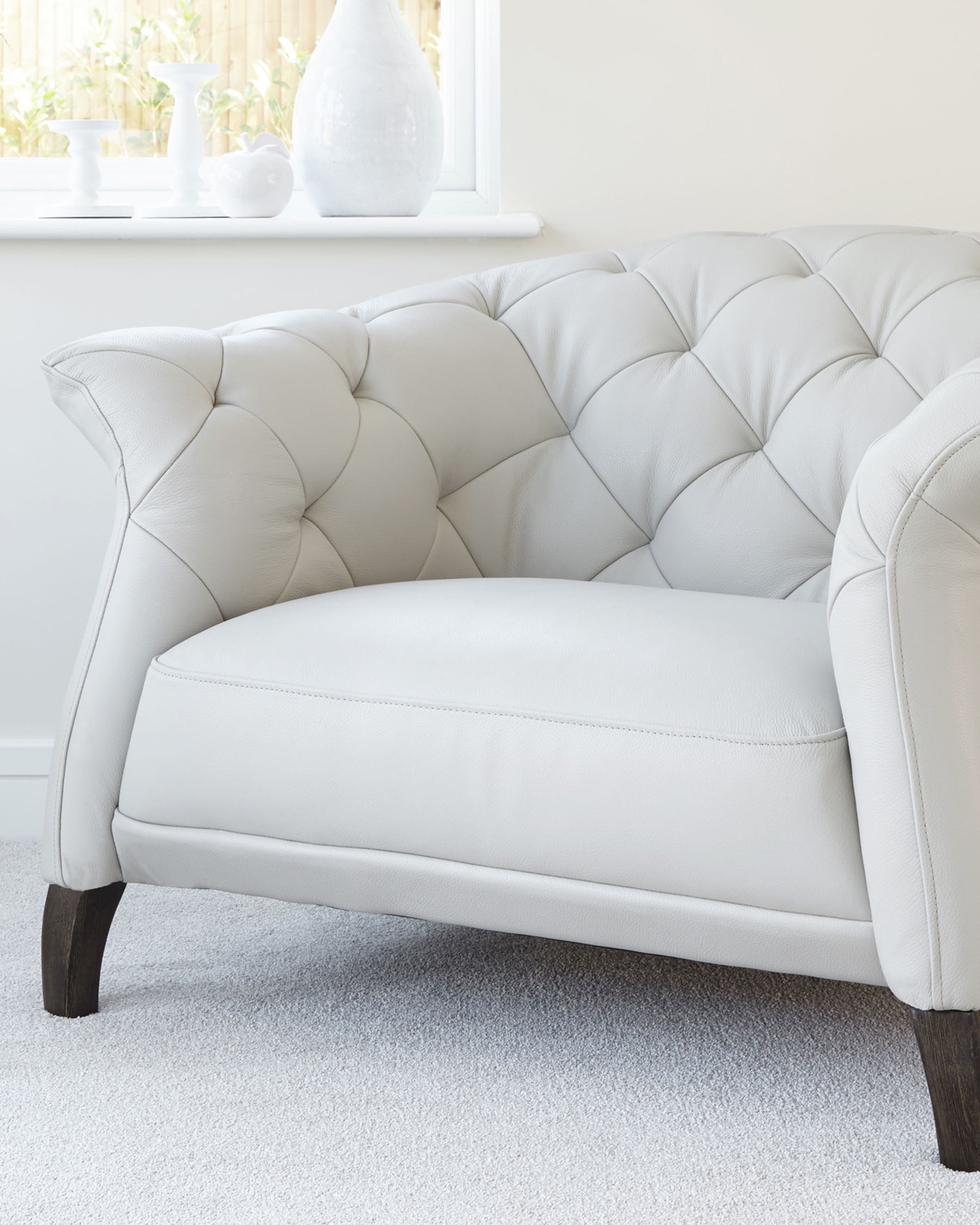 Elegant cream-colored leather loveseat with tufted back cushioning and plush seat. The seat has rounded arms and sits on dark wooden legs, providing a classic touch to a contemporary design. Positioned in a bright room near a window with natural light, complemented by neutral decor on the window sill.