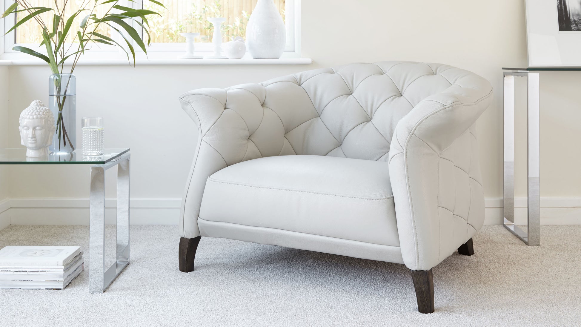 Elegant white tufted wingback chair with tapered dark wooden legs, paired with a modern glass side table with metallic legs featuring decor items.