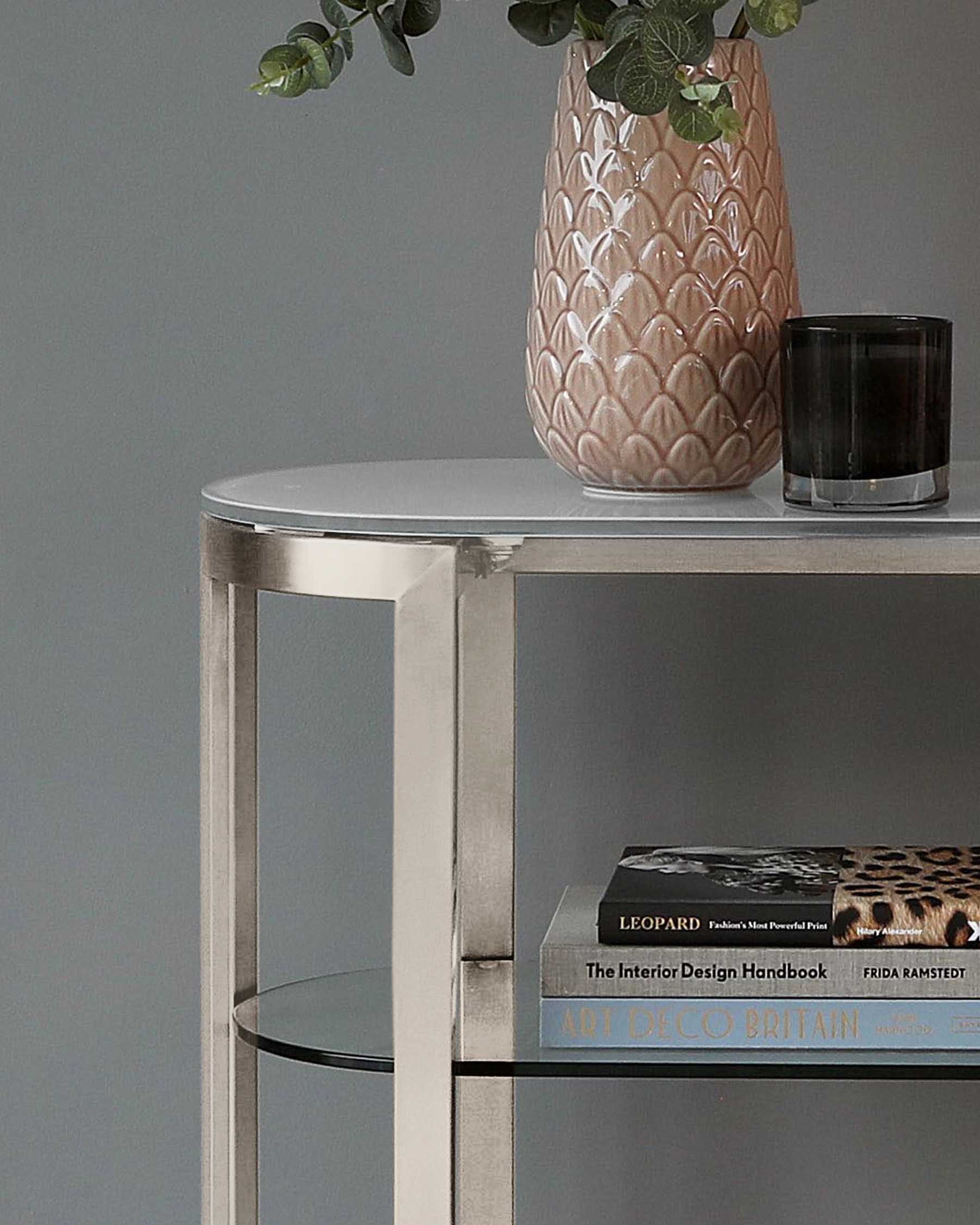 A contemporary round side table with a polished metallic finish and a mirrored top surface, featuring an open cylindrical structure and a secondary mirrored shelf holding decorative books.