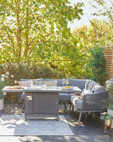 Outdoor patio furniture featuring a modern grey corner sofa with plush cushions and a matching dining table with a grey tabletop and a built-in gas fire pit. The set is complemented by a patterned floor rug and potted plants, creating a cosy outdoor living space.