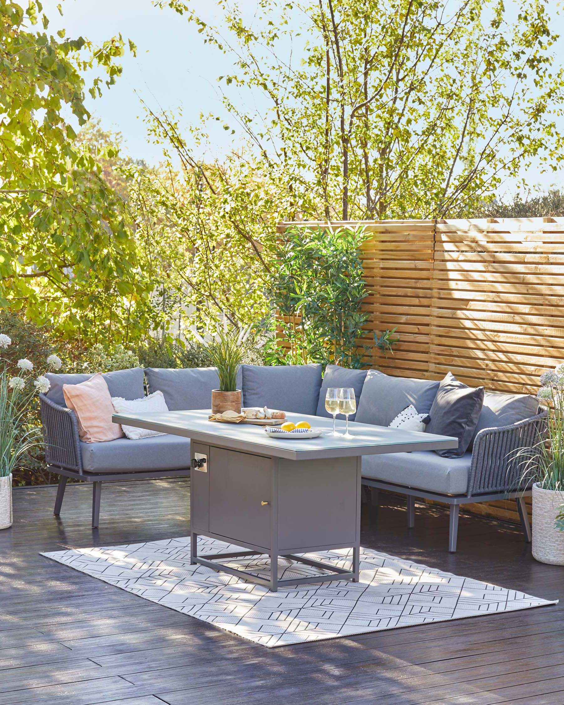 Outdoor furniture set comprising a rectangular dining table with a sleek grey finish and a matching bench and chairs with dark grey woven wicker detailing and light grey cushioning. A simple geometric patterned rug lies underneath, set against a warm wooden deck background, enveloped by lush greenery.