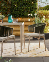 A light-colored outdoor table with two green, woven chairs featuring beige cushions, all set on a soft, patterned rug.