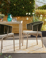 Outdoor furniture set featuring a modern, minimalist white square table with sleek lines and two contemporary-style chairs with dark green vertical strap backs and beige seat cushions. The set is displayed on a beige and cream patterned outdoor rug, with lush greenery and wooden fence panels in the background, accentuated by small string lights. A teal glass pitcher and clear drinking glasses adorn the table top, creating a welcoming ambiance for outdoor dining or leisure.