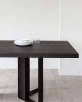 Modern minimalist dark wood dining table with a thick tabletop and sturdy rectangular legs, set against a neutral background with light wood flooring. Display includes a stack of white plates and clear glasses on top.