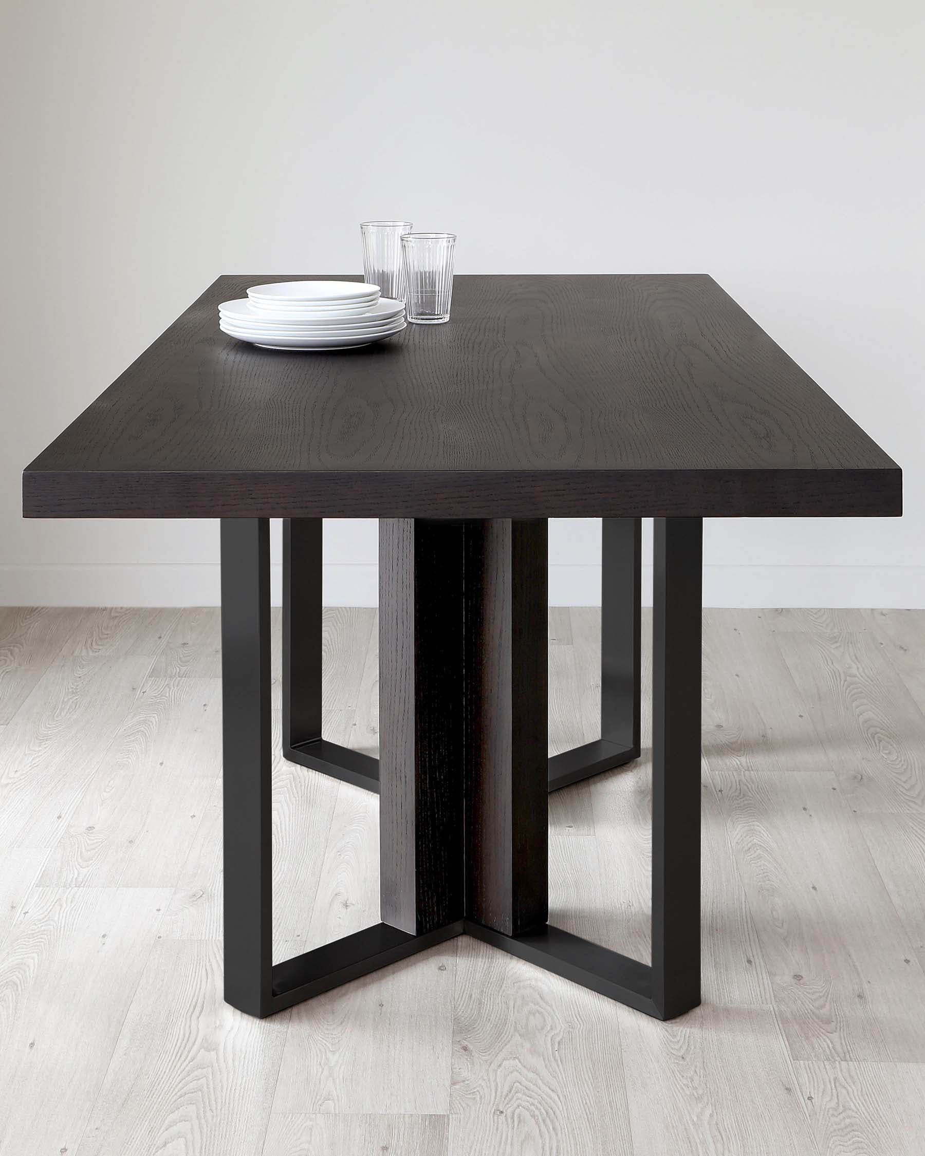 A contemporary dining table with a dark wood finish and a robust, double pedestal base featuring angular metal supports. The tabletop, adorned with a minimalistic arrangement of white plates and clear glasses, showcases the natural wood grain pattern.