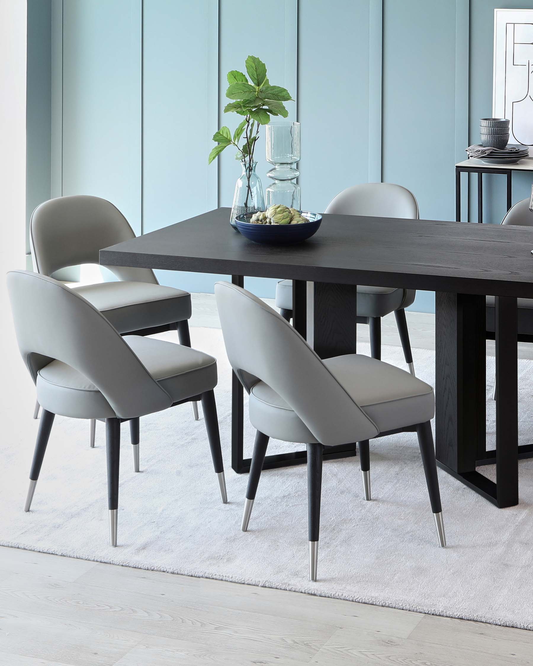 Modern dining room furniture set with a sleek, dark wooden table and six light grey upholstered chairs featuring black tapered legs with metallic tips. The table is adorned with a glass vase containing greenery and a blue decorative bowl. The set is placed on a light-coloured area rug over hardwood flooring, against a backdrop of pale blue walls with minimalist artwork and a console table with a stack of dishes.