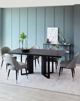 Modern dining room furniture set featuring a rectangular black wooden table with thick, vertical slab legs, paired with four sleek grey upholstered chairs with black wooden frames and angled, tapered metal legs. A minimalist two-tiered black shelving unit stands to the right holding decorative items. The set is accentuated by a soft white area rug on light wooden flooring.