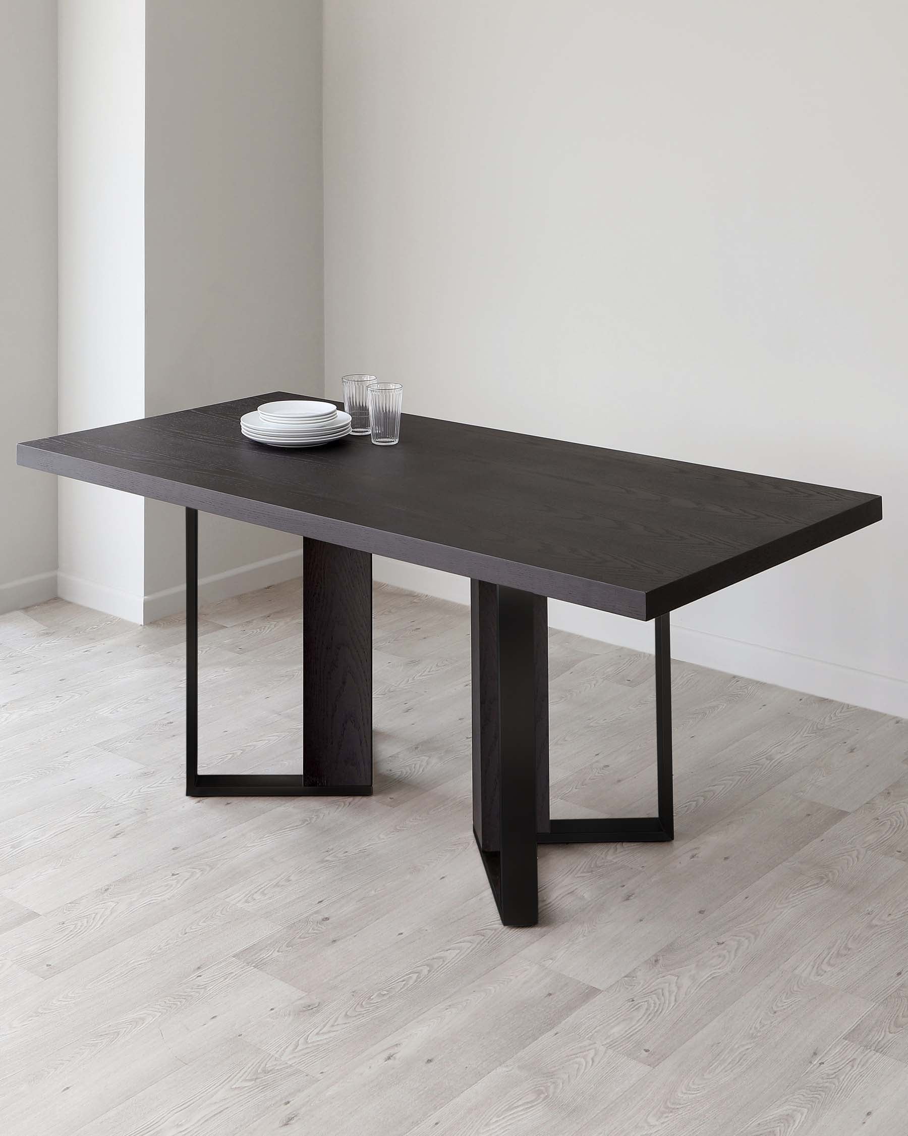 A modern dark wooden dining table with a rectangular top and two sturdy, inverted V-shaped legs, featuring a minimalistic design. On the table, there is a simple setting of white dinner plates stacked together and two clear drinking glasses. The table is placed on a light hardwood floor against a neutral background, showcasing its sleek silhouette.