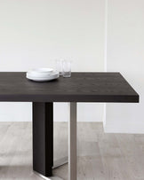 Modern minimalist dark wood dining table with a sturdy rectangular top and a bold single pedestal base, accented with a metallic strip at the foot. The table is subtly adorned with a stack of white plates and clear drinking glasses, set against a neutral backdrop, highlighting the table's sleek design.