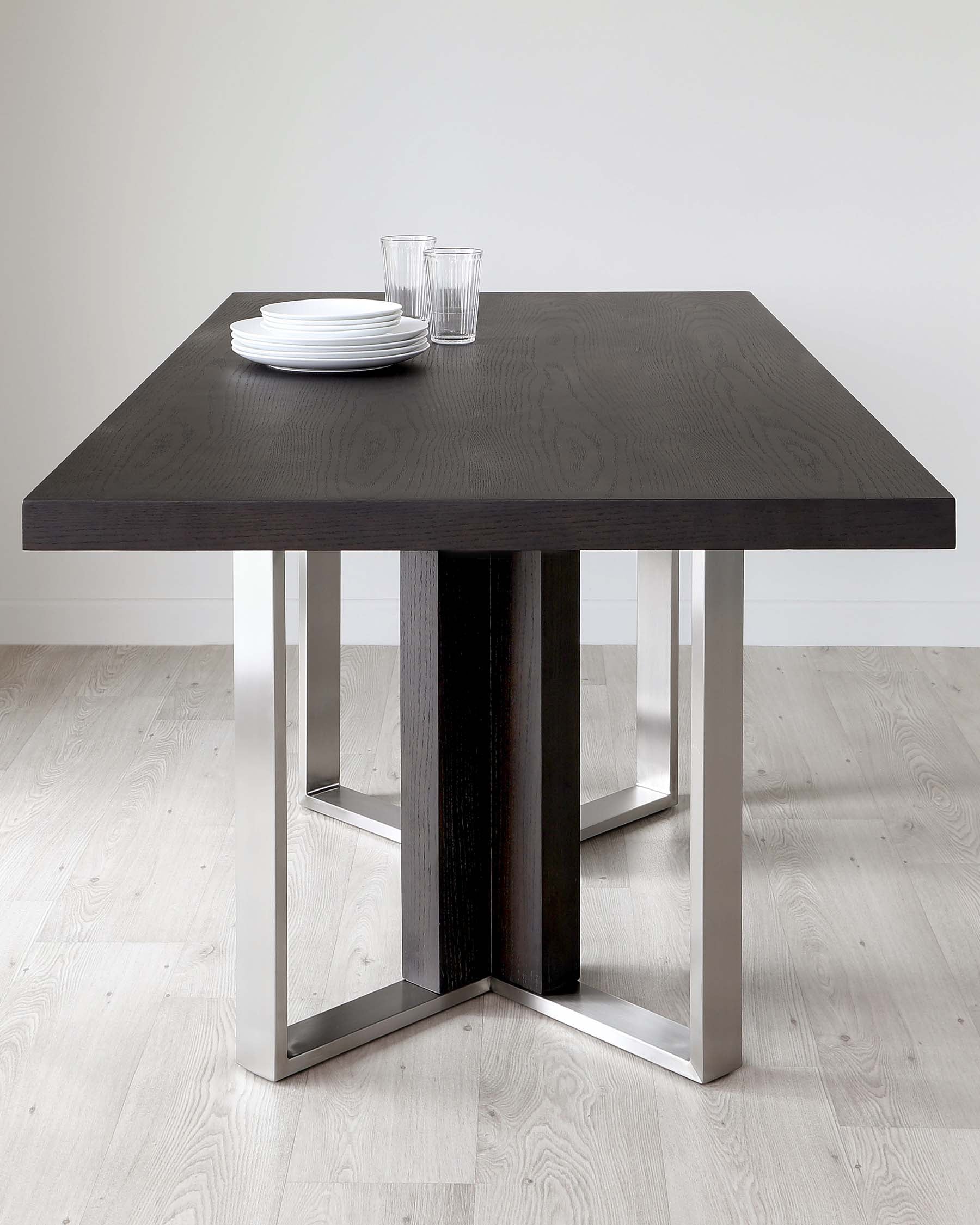 Modern square dining table with a textured dark wood finish and a unique geometric base featuring wood and chrome elements. On top of the table are a stack of white plates and two clear glasses, suggesting readiness for a casual meal.