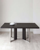 Modern minimalist dark wood dining table with sturdy rectangular top and two prominent leg structures, each featuring a pair of vertical panels connected by a horizontal chrome bar at the base, on a light hardwood floor against a plain white wall, adorned with a simple white dinnerware set and clear drinking glasses.