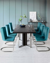 A modern dining set featuring a dark wood rectangular table with a robust, double-pedestal base and six luxurious teal upholstered chairs with sleek, curved metal legs. A coordinating sideboard with decor is placed against the wall.