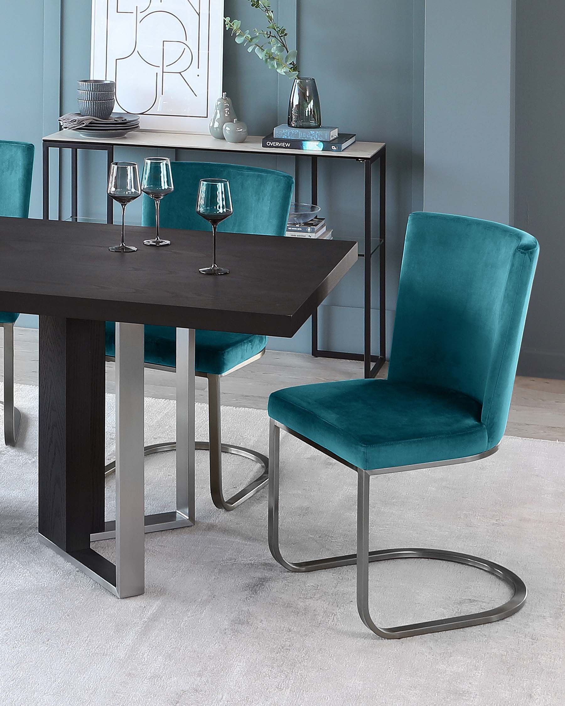 Modern dining room furniture set featuring a dark wood table with solid rectangular top and sturdy block legs, paired with luxurious teal velvet upholstered chairs with unique cantilevered metal bases. A coordinating sideboard with open shelving displays decorative items, enhancing the contemporary design aesthetic.