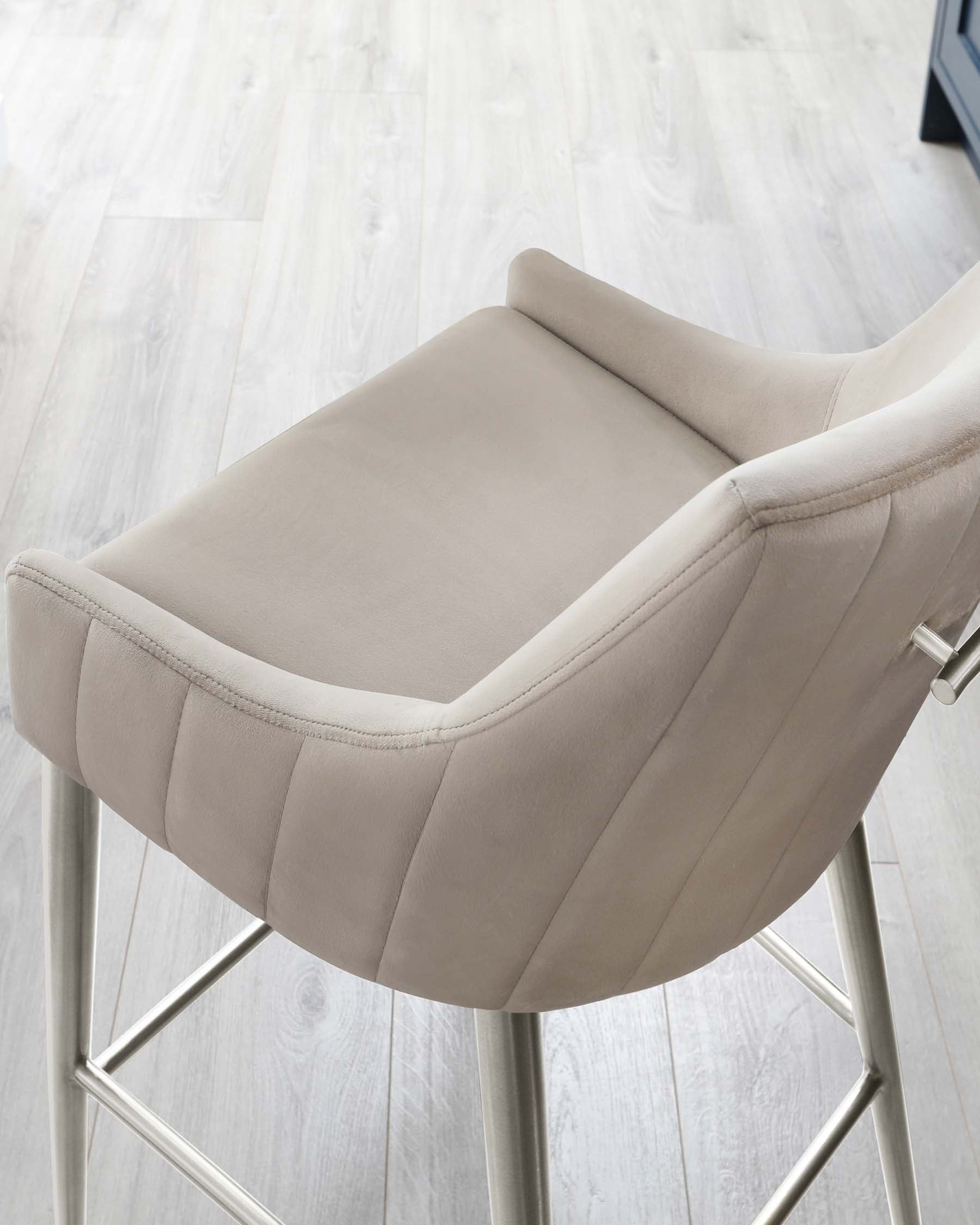 Modern light grey upholstered bar stool with a curved backrest and channel tufting details. Features a sleek metallic base with a footrest, set against a light wood laminate flooring background.