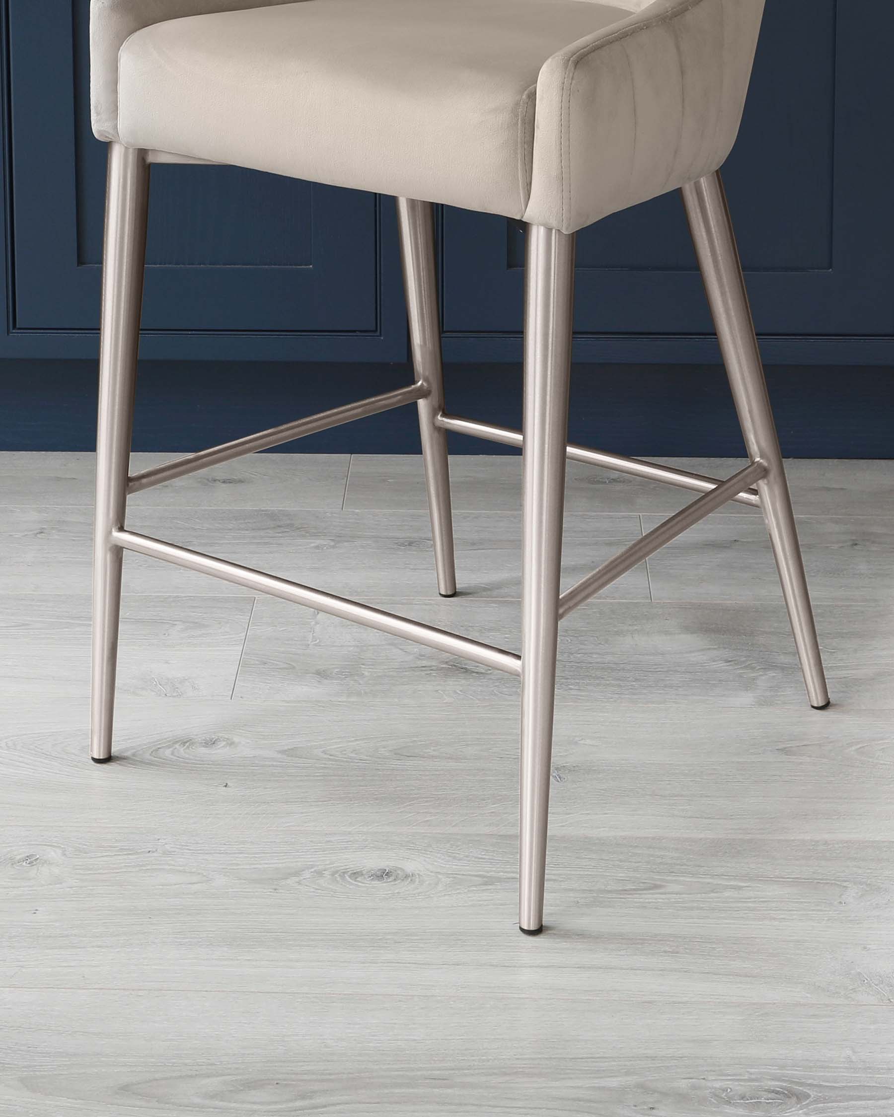 Elegant modern bar stool with a plush, light beige upholstered seat and backrest, complemented by sleek, brushed metallic legs in a geometric pattern with footrests for added comfort.