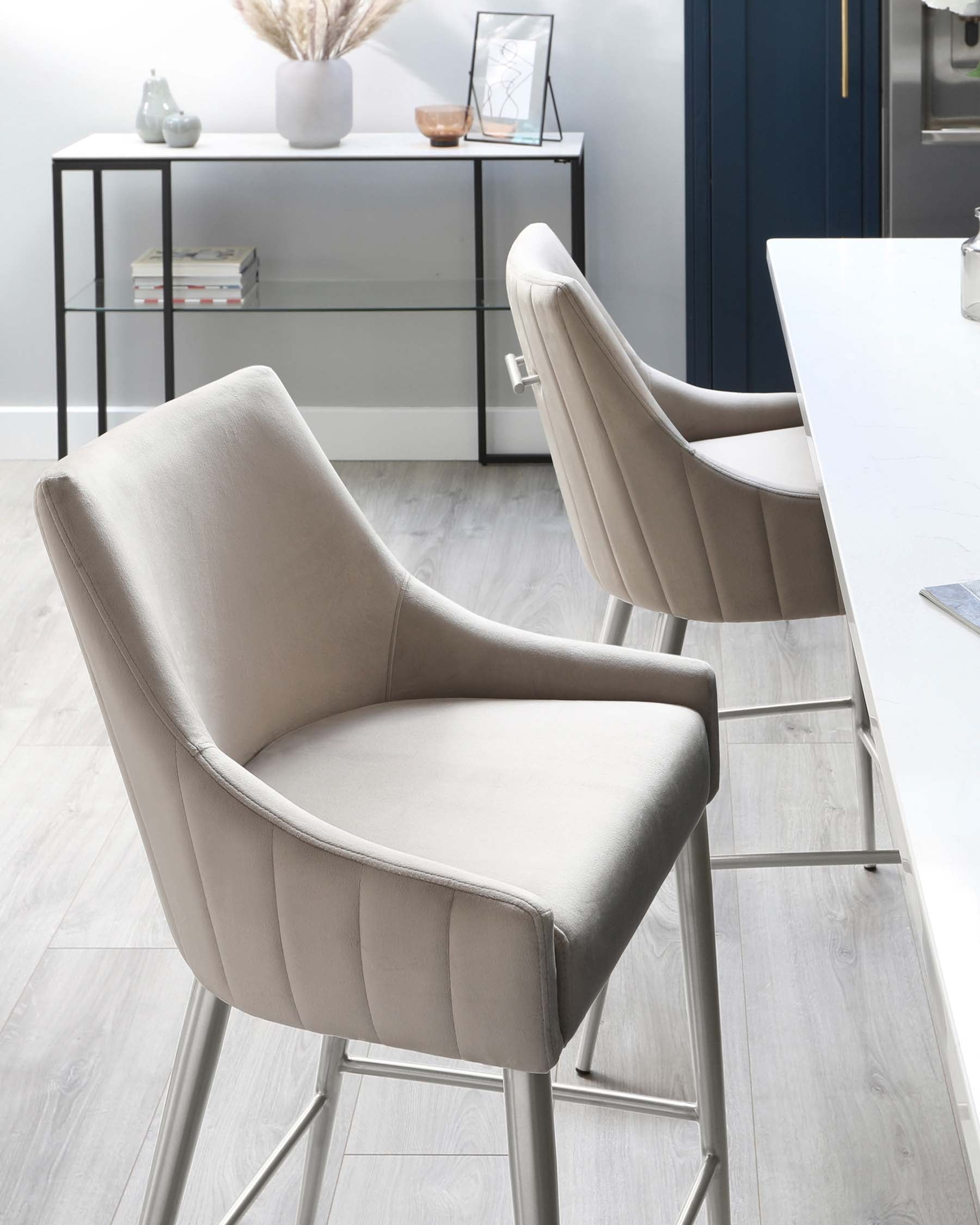 Two modern upholstered bar chairs with a smooth, light grey fabric cover and sleek metal legs in the foreground. A minimalist black metal console table with a glass shelf in the background, adorned with decorative items including a vase, frames, and books.