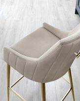 Elegant modern barstool with a curved, cushioned backrest and a comfortable, padded seat in a neutral beige upholstery. The chair features sleek, gold-coloured metal legs that have a geometric flair, providing a sophisticated and stylish look suitable for contemporary kitchen or bar settings.