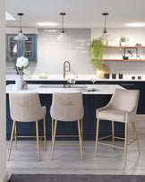 Modern kitchen with three cream upholstered bar stools with low backrest and sleek golden metal legs, positioned at a dark navy kitchen island with a white countertop. A plush armchair with matching cream upholstery and gold legs stands to the side, complementing the sophisticated colour palette of the space.