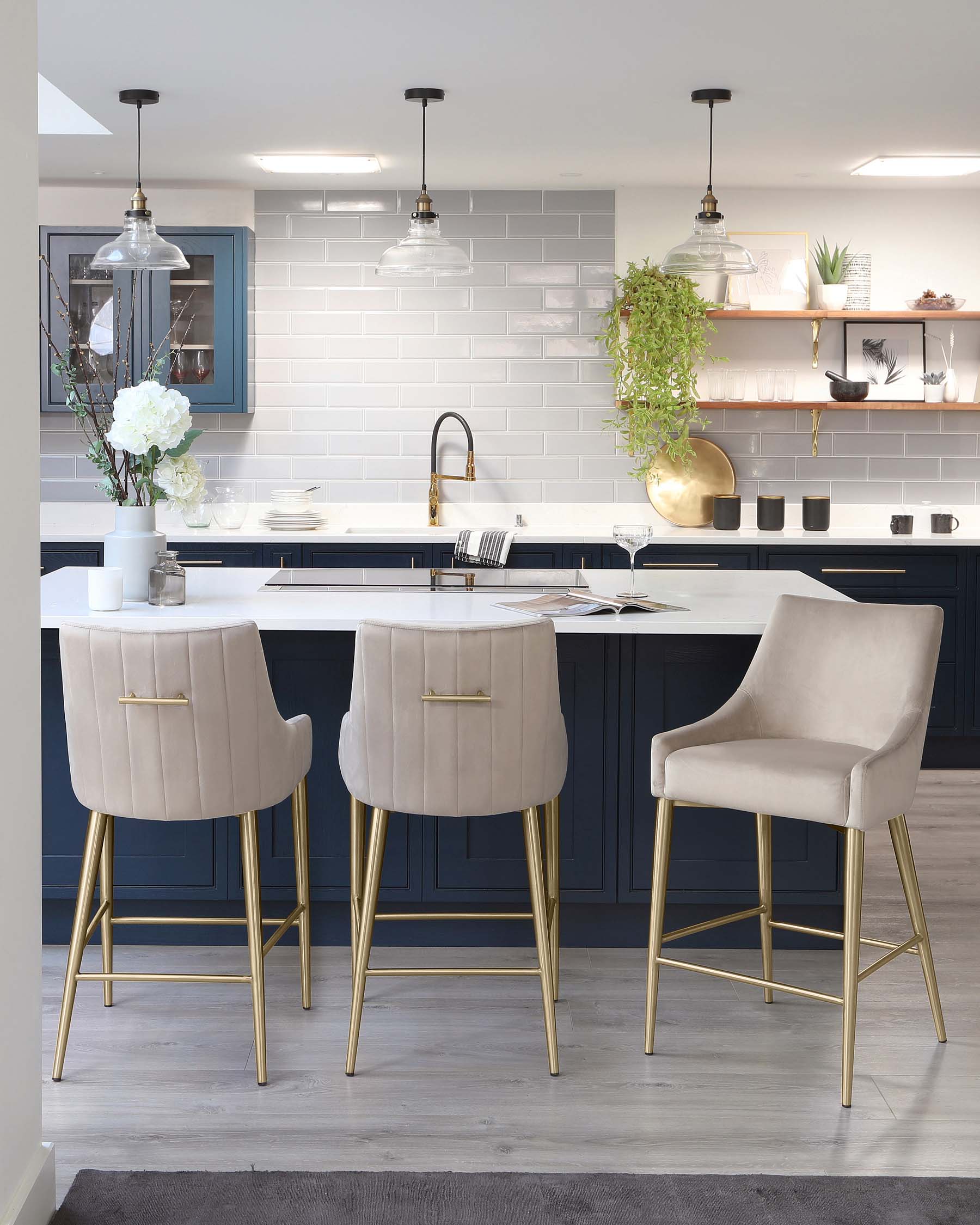 Modern kitchen with three cream upholstered bar stools with low backrest and sleek golden metal legs, positioned at a dark navy kitchen island with a white countertop. A plush armchair with matching cream upholstery and gold legs stands to the side, complementing the sophisticated colour palette of the space.