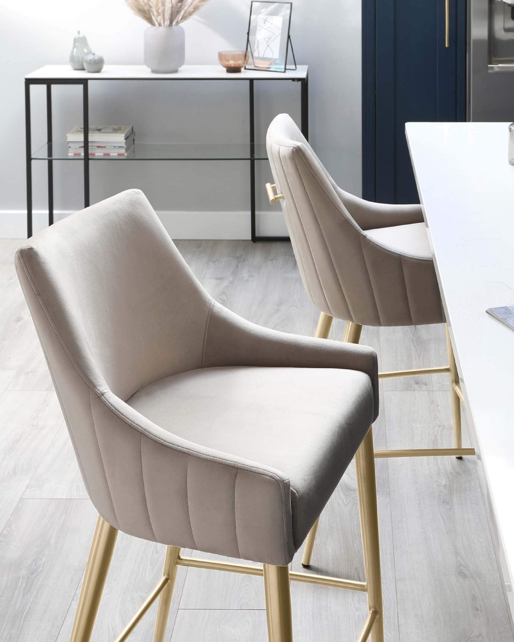 Elegant modern dining chairs with beige upholstery and gold metal legs in front of a sleek, white marble table, with a minimalist black metal console table featuring glass shelves in the background.