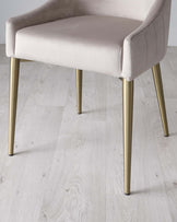 Elegant modern chair with plush light beige upholstery and channel tufting on the backrest, seated on tapered metallic gold legs. Set against a light wood floor background.