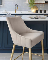 Elegant modern bar stool with a beige upholstered seat and backrest, featuring a channel tufting design, set atop a sleek metallic gold-finished frame with slender legs and a geometric footrest.