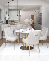 Elegant dining room set featuring a round, white, marble-topped table with a brass-coloured metal base, surrounded by six plush, beige upholstered chairs with vertical stitching and slender, tapered, brass-coloured metal legs. A white rug underlines the set, offering a chic and contemporary appeal.