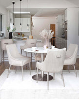 A modern dining area with a round white table and four complementary plush upholstered chairs in a soft beige colour, featuring high backs and slender metal legs. A matching sideboard with a flat surface holding decorative items is visible in the background. The setting includes a muted white rug under the dining set, enhancing the contemporary and clean aesthetic of the space.