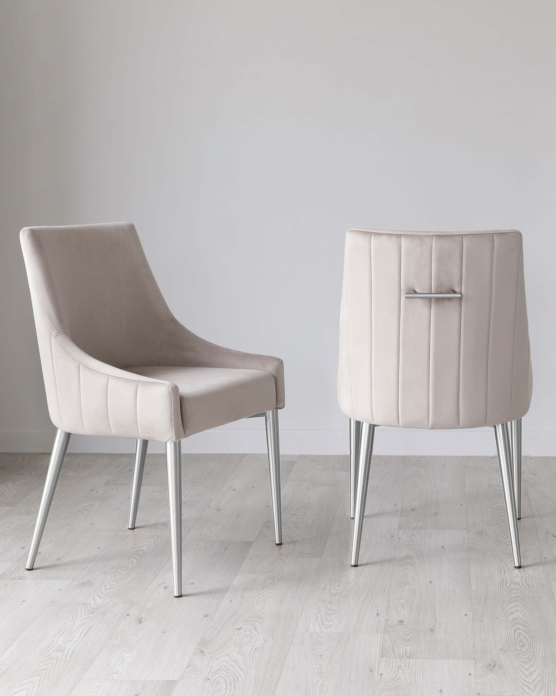 Two modern chairs with beige upholstery and sleek silver metal legs, featuring a smooth curved silhouette and linear stitching detail, displayed on a light hardwood floor against a neutral backdrop.