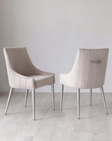 Two modern upholstered dining chairs with sleek, silver metal legs, featuring a gently curved backrest and comfortable seat cushion in an elegant light taupe fabric. The design includes subtle horizontal stitching on the backrest, adding a touch of sophistication. Chairs are positioned on a light hardwood floor against a plain white background.