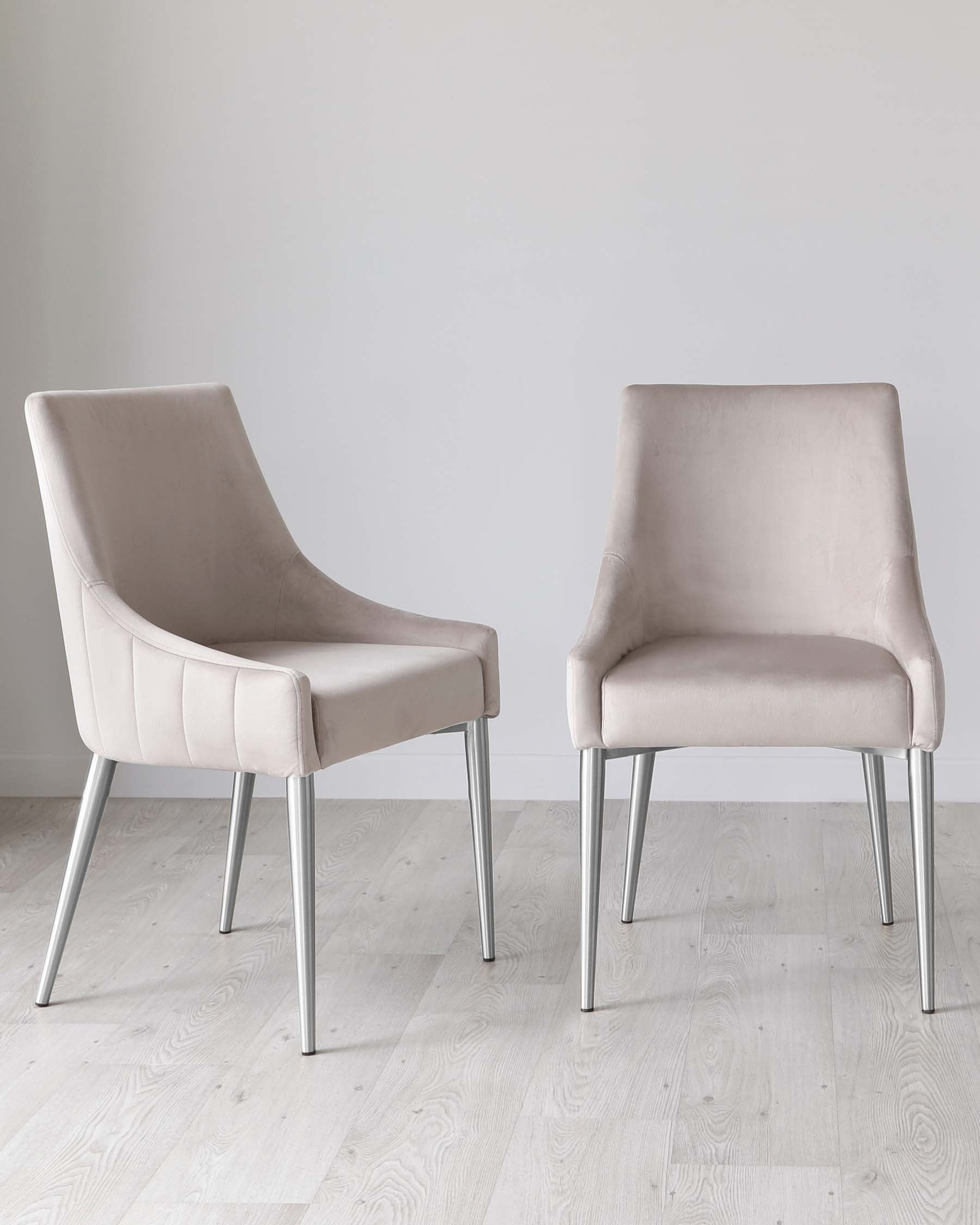 Two contemporary dining chairs with beige upholstery and sleek, silver metal legs, standing on a light wooden floor against a white wall.