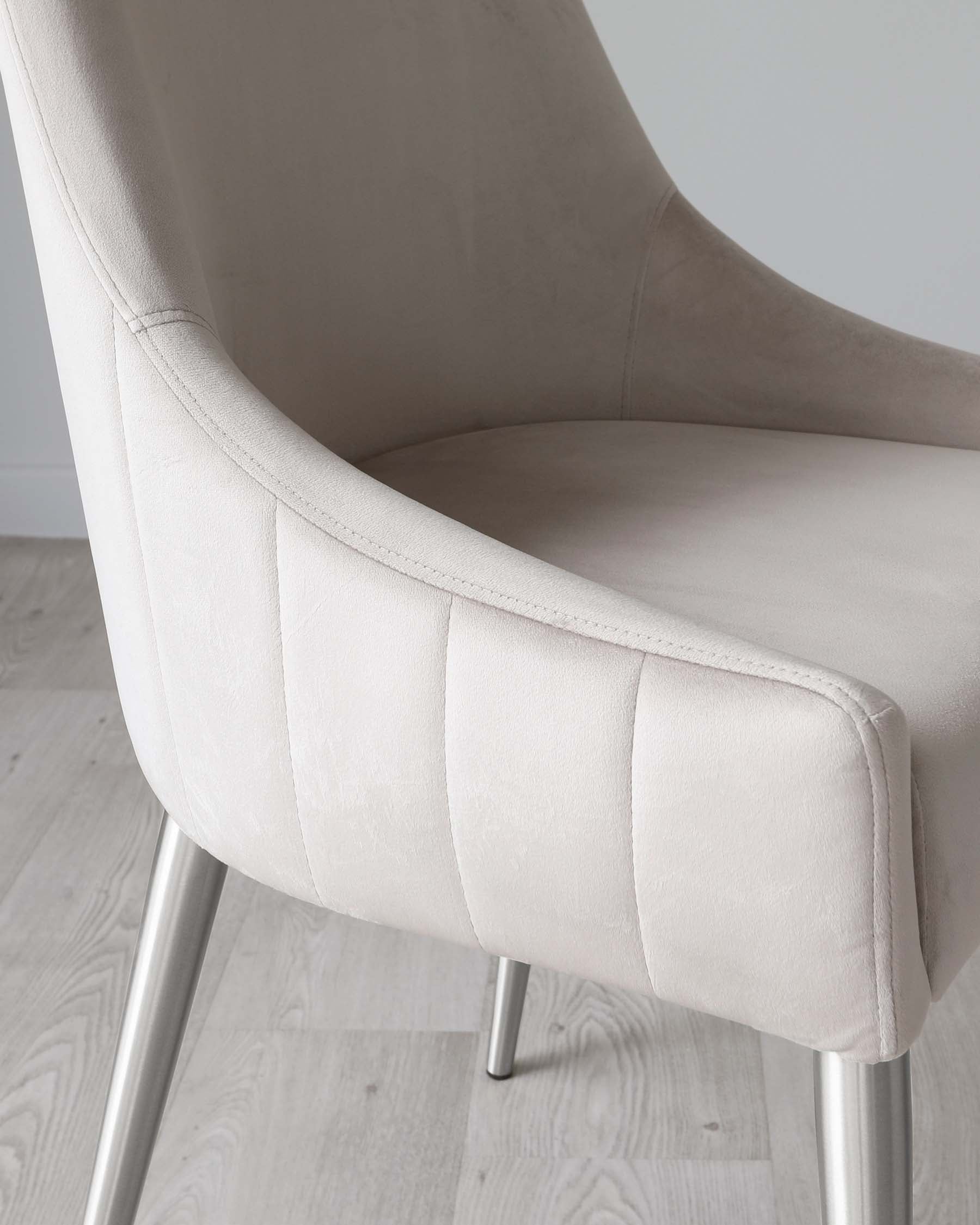 Modern light beige upholstered chair with a smooth curvilinear shape and channel tufting detail on the seat back. It features a slightly reclined backrest for added comfort, rounded armrests, and is supported by sleek, tapered metallic legs. The chair stands on a light wood flooring against a clean white wall background.