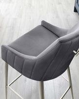 Modern grey upholstered bar stool with channel tufting on the backrest and cushioned seat, featuring sleek metal legs with a geometric design and a footrest.