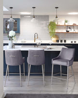 Three modern bar stools with sleek grey upholstery and slender metallic legs positioned around a kitchen island.