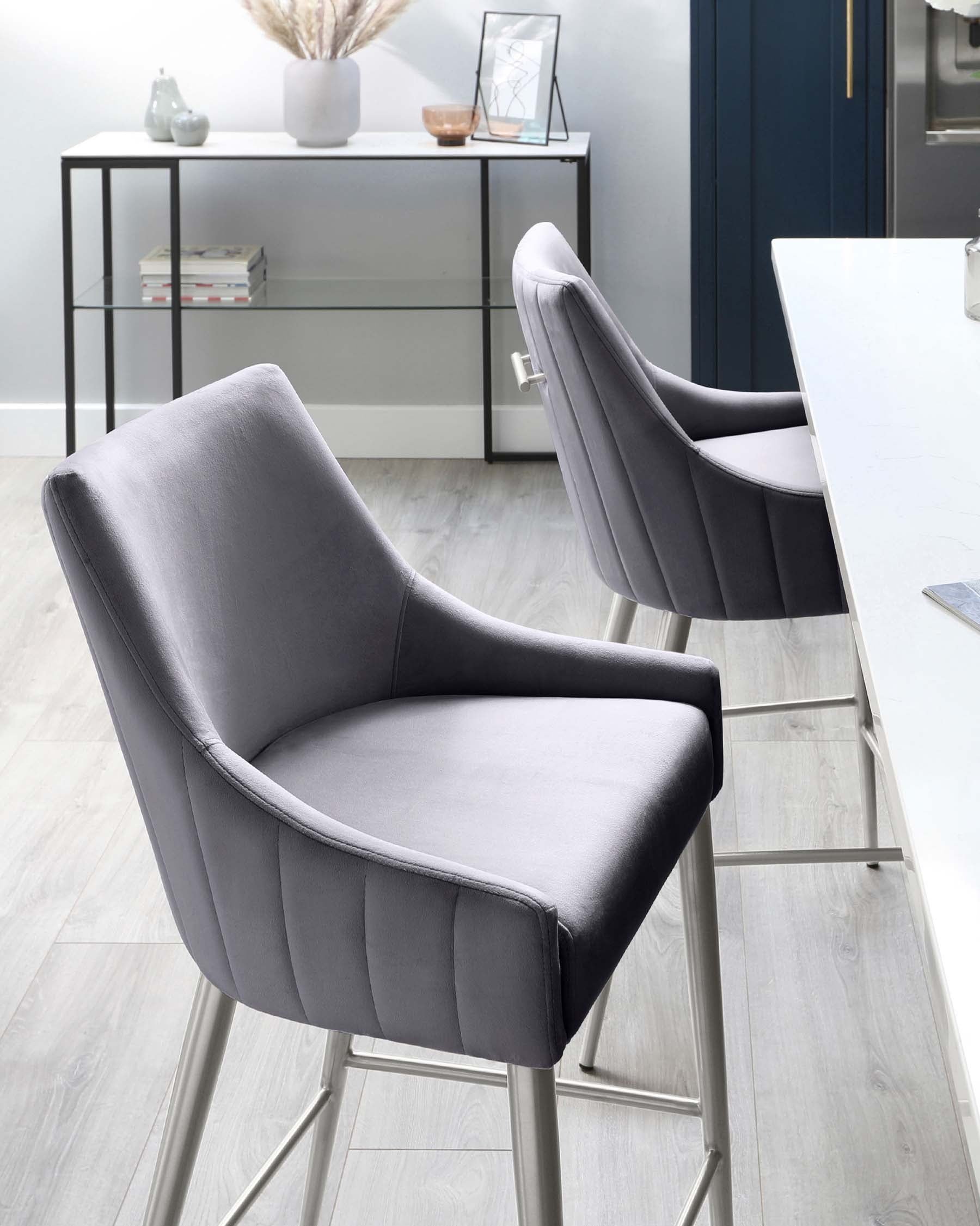 Two elegant grey upholstered bar chairs with a smooth finish and vertical stitching detail on the back. A sleek black metal console table with a glass top and two lower shelves displays decorative vases and frame in a modern kitchen setting.