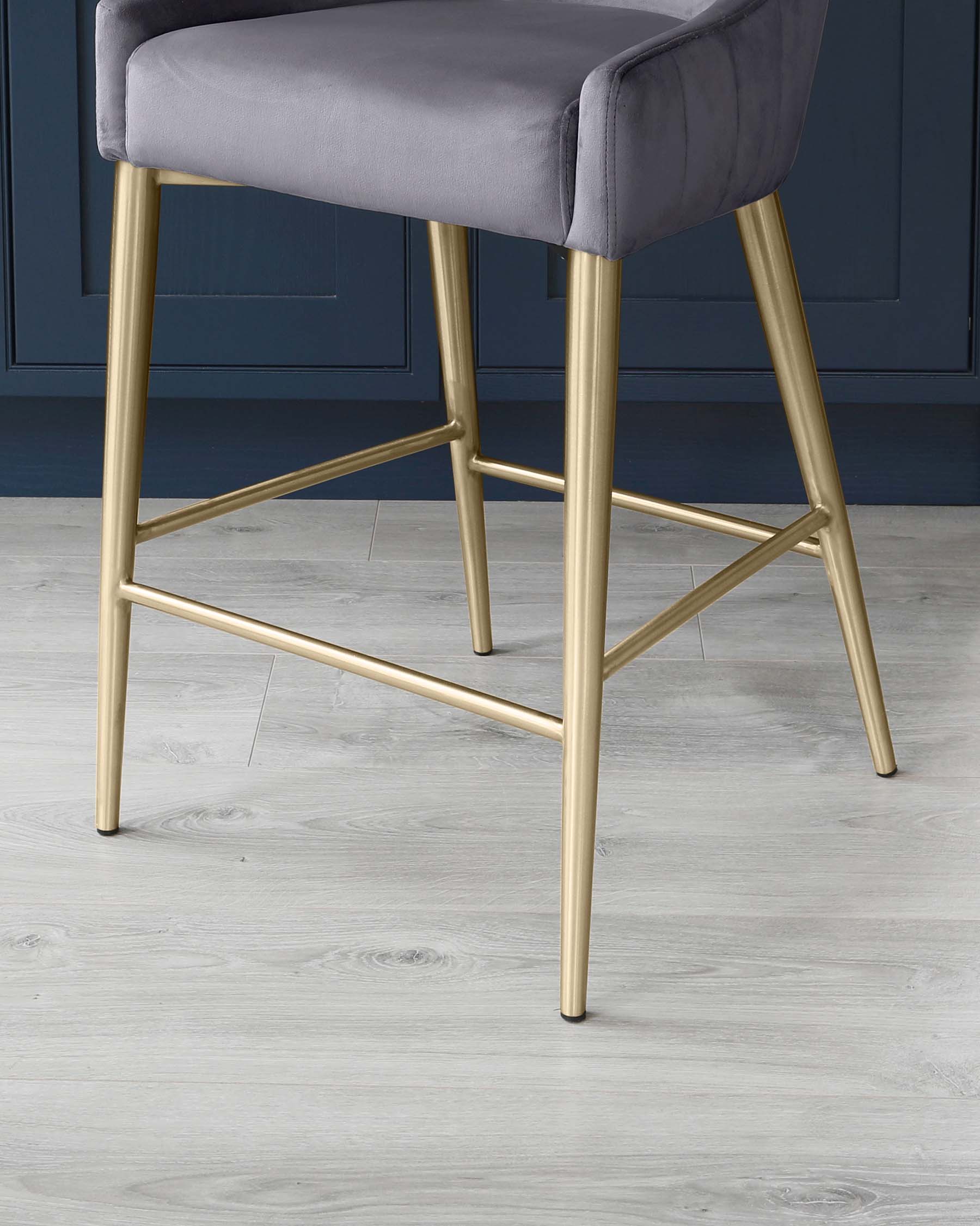 Modern grey upholstered bar stool with sleek gold metal legs on a light wood floor, against a deep blue wall panel background.