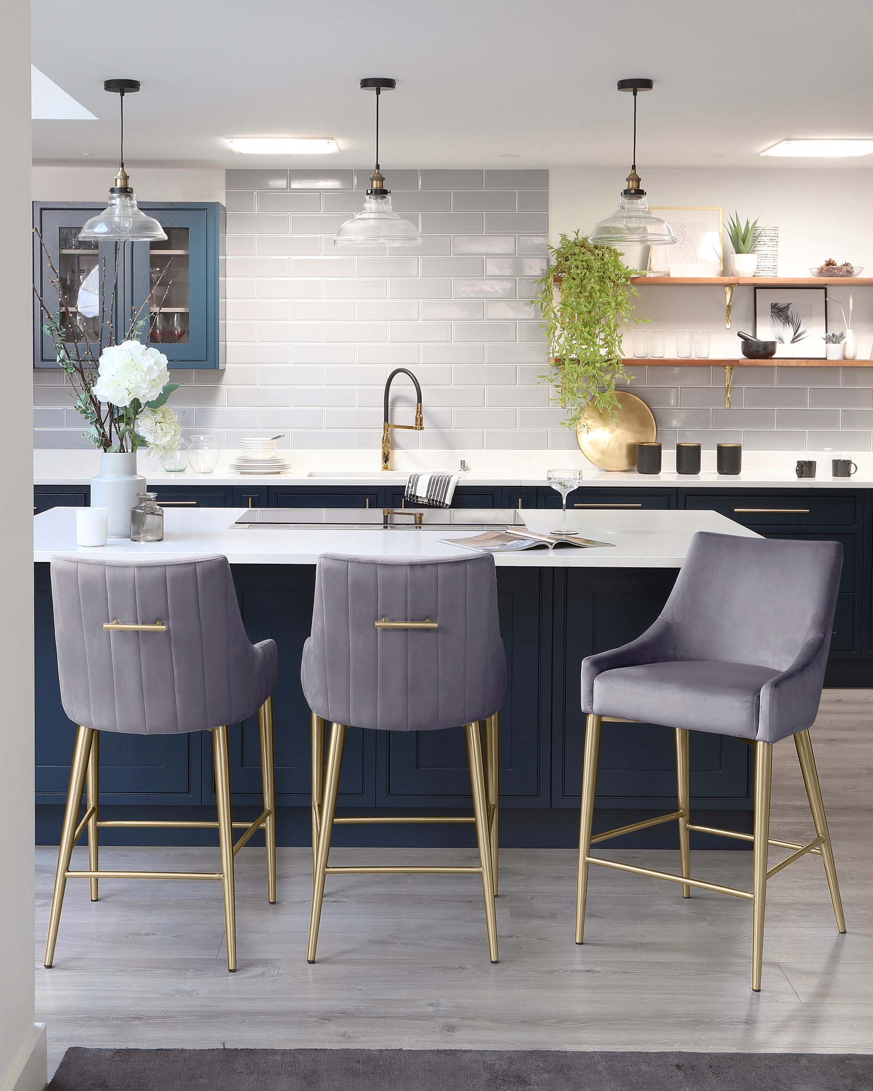 Three modern bar stools with plush, grey upholstered seats and backs, featuring subtle tufted detailing. Each stool stands on elegantly slender, brass-finished metal legs, which include footrests for added comfort. The furniture exudes a blend of contemporary design with a touch of luxury, perfect to enhance a stylish kitchen island or bar area.